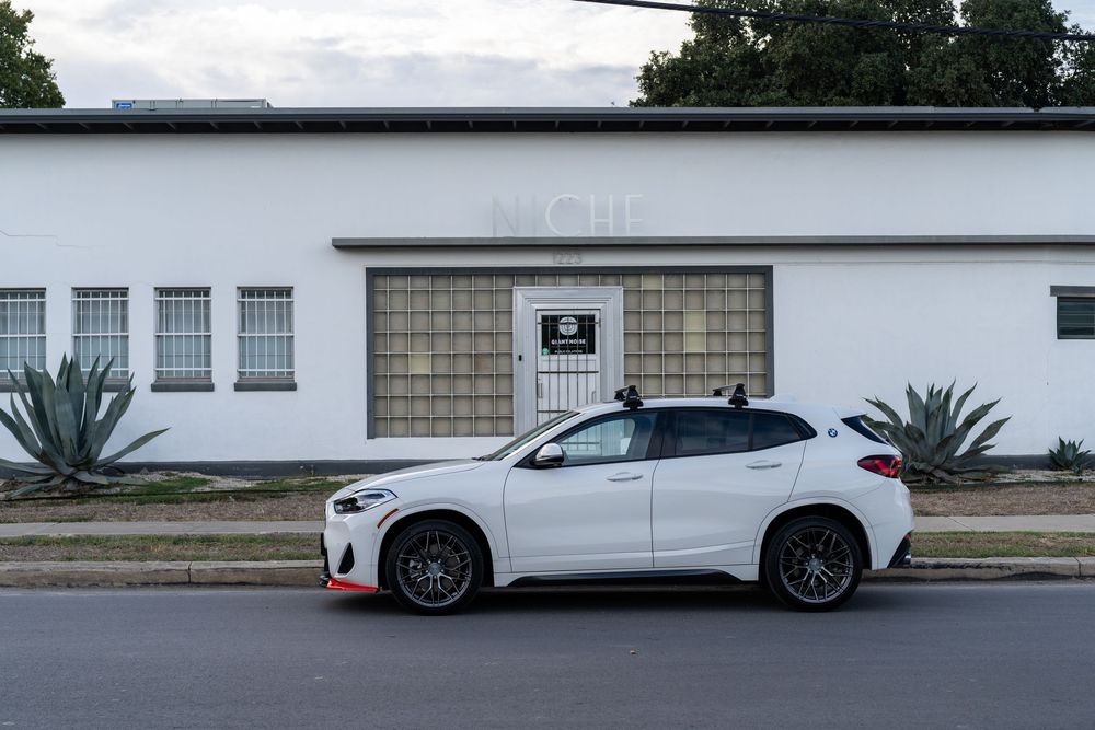 A white Porsche Cayenne SUV parked in front of a modern building with palm trees visible in the background.