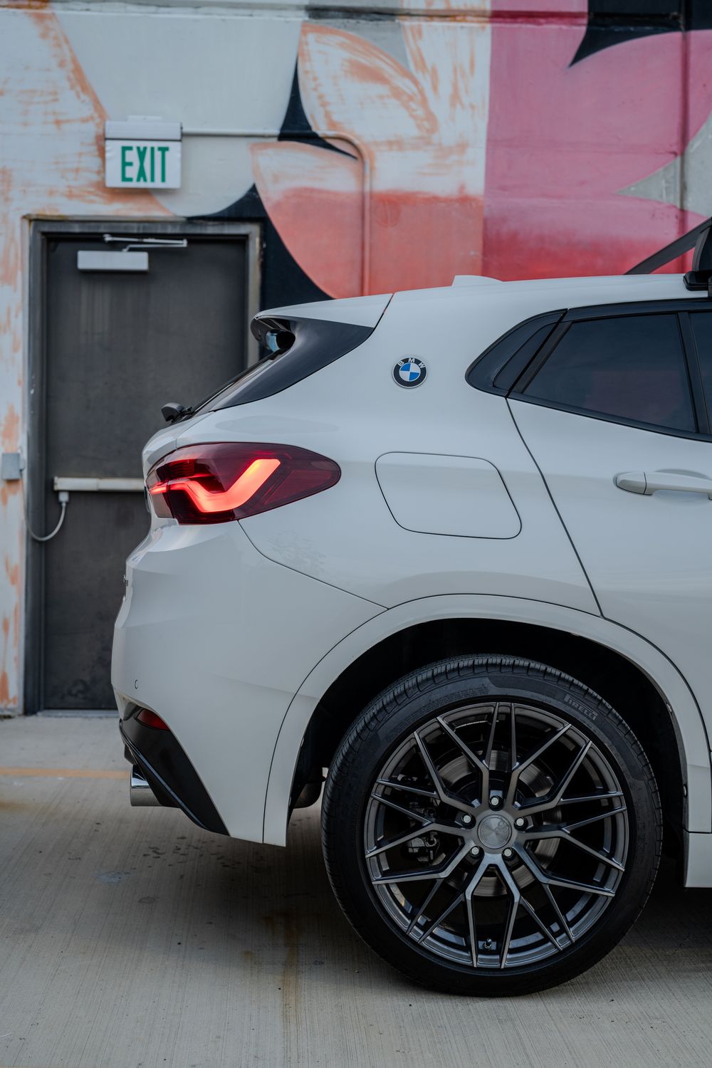 Close-up view of a white BMW's rear quarter panel and black alloy wheel against a pink wall with an exit sign.
