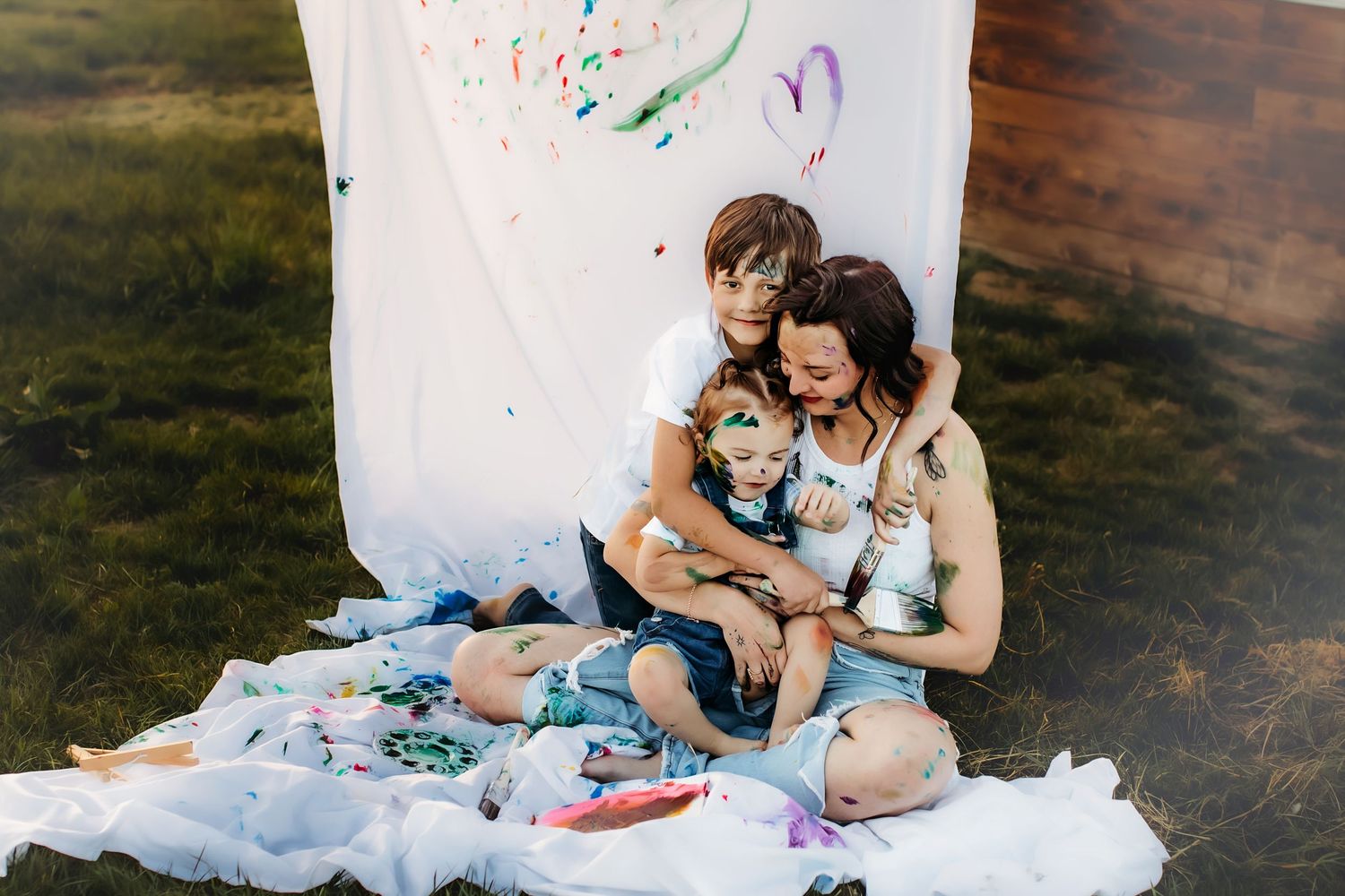 Family snuggling together on white blanket with colorful paint splatters during outdoor photo session.