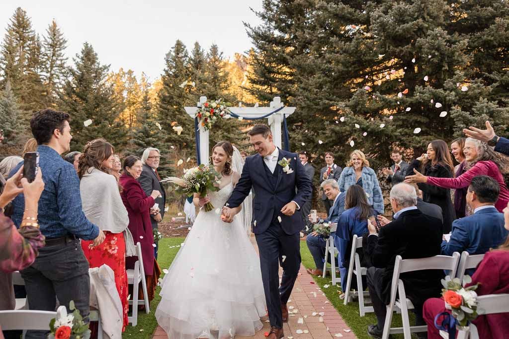 Wedding ceremony guests gather outdoors surrounded by tall evergreen trees during a golden sunset hour.
