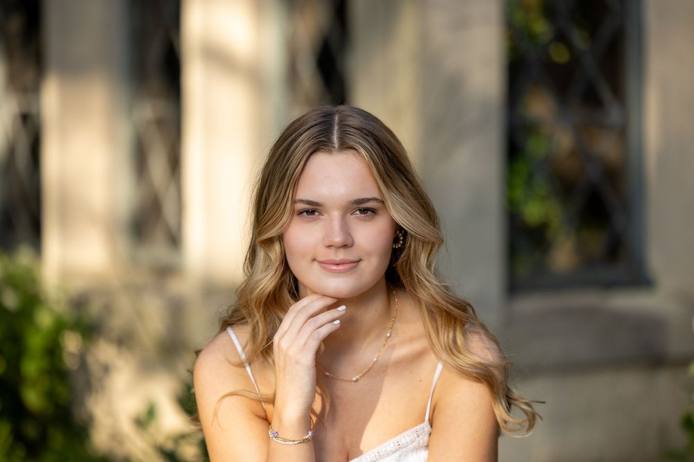 A blonde model in a peach-colored top poses outdoors with natural lighting and a soft smile.