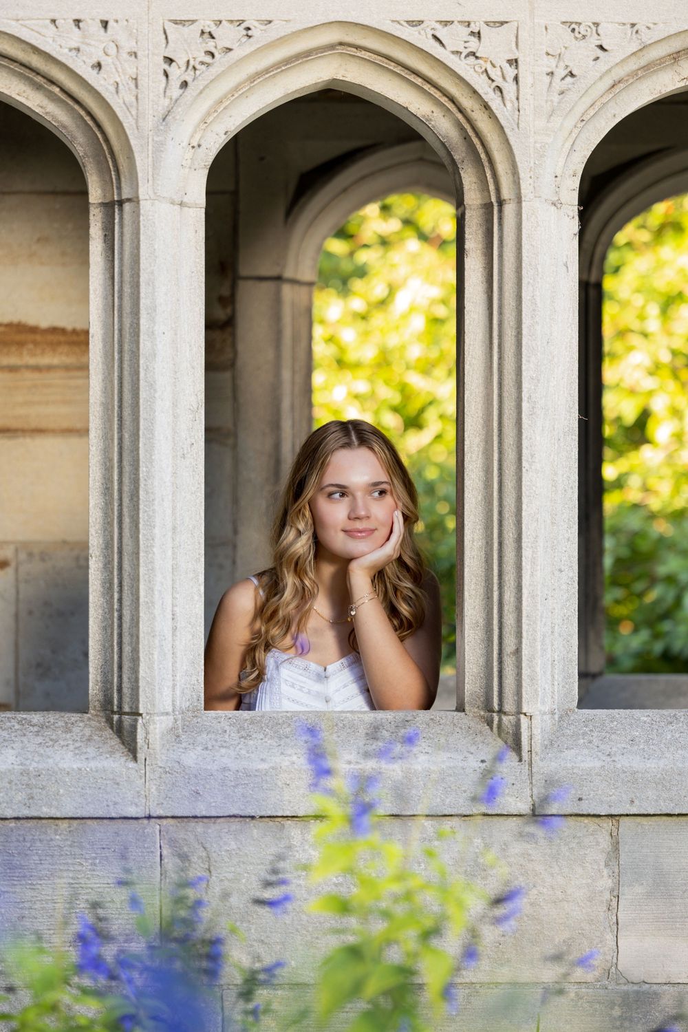 A person in a white dress poses thoughtfully in an ornate stone archway with blurred yellow and purple flowers in foreground.