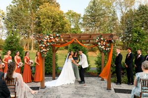 Wedding party gathers under wooden pergola in autumn garden setting with orange and white floral decorations.