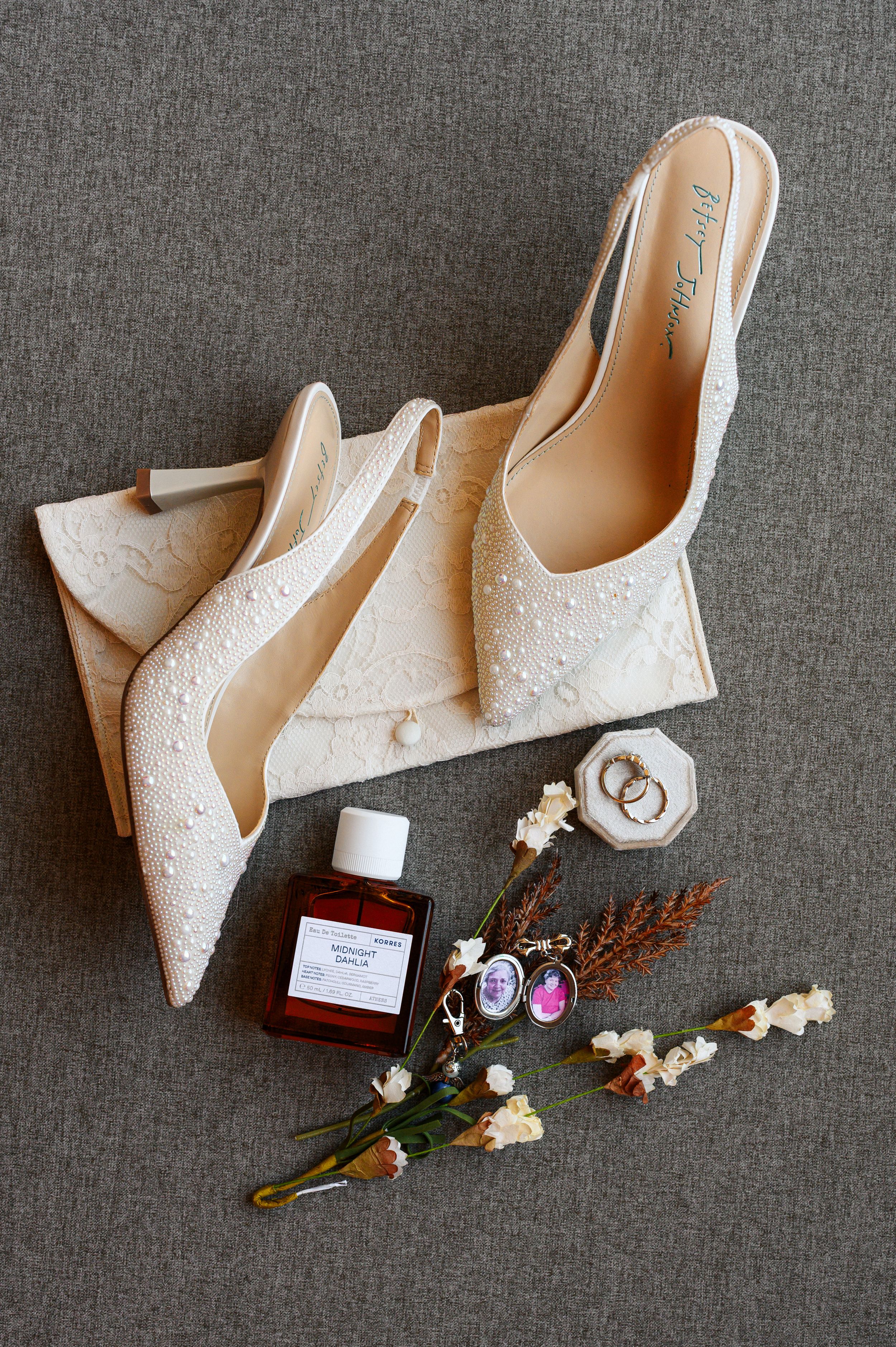 White bridal shoes with delicate beading displayed alongside wedding rings and floral decorations on a gray background.