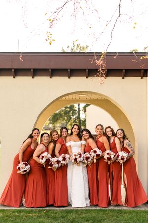Wedding party in matching rust-colored bridesmaid dresses and white bridal gown pose under arched doorway with floral bouquets.