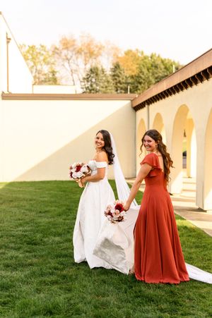 Two women in formal dresses, one in white and one in rust-colored, stand on grass holding red floral bouquets.