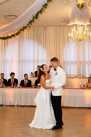 A couple shares their first dance in an elegant white ballroom decorated with chandeliers and draped fabric.