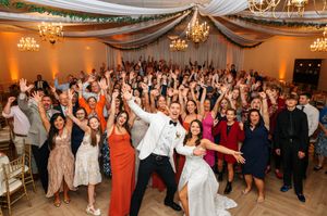 Large group of wedding guests dancing and celebrating together on a dance floor under elegant chandeliers.