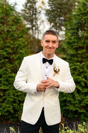 A person in a white tuxedo jacket and black bow tie poses playfully in front of evergreen trees at an outdoor event.