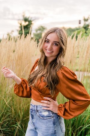 Person in rust-colored top and denim jeans poses playfully in tall grass field during golden hour.