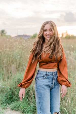 A person in a rust-colored blouse and blue jeans poses playfully in a sunlit meadow during golden hour.