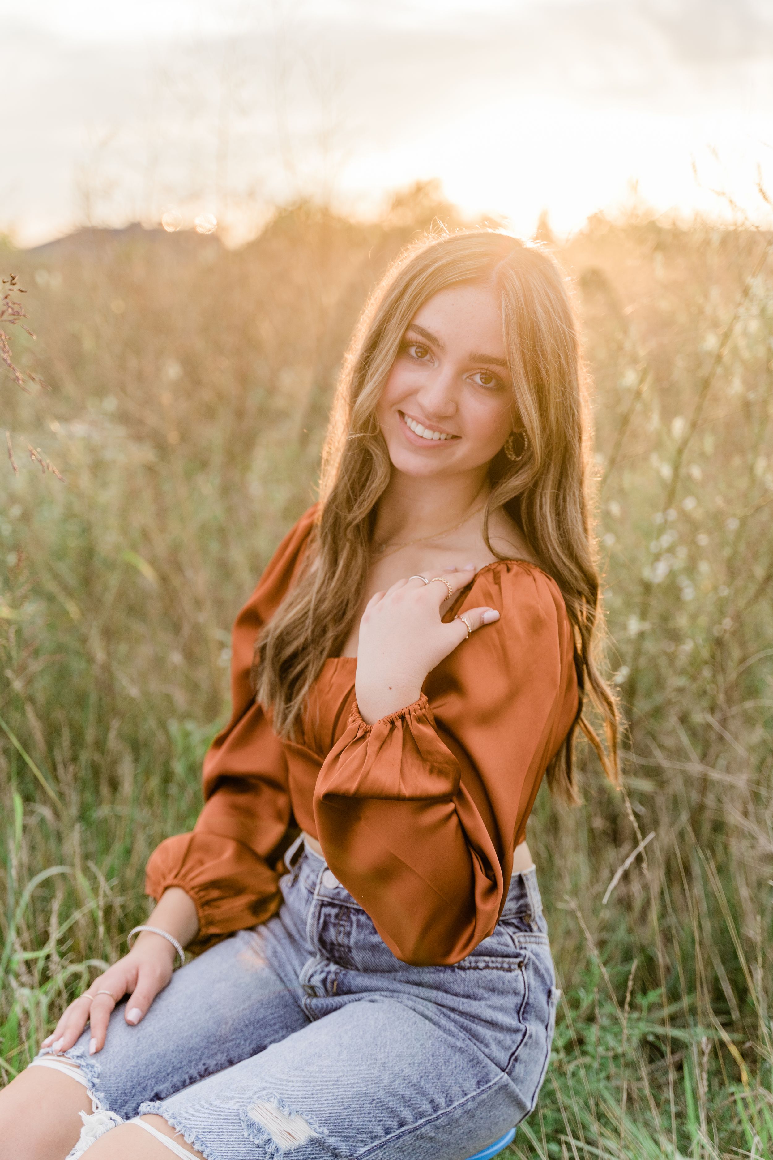 Person in orange blouse and jeans sits casually in sunlit meadow with tall grass swaying in background.