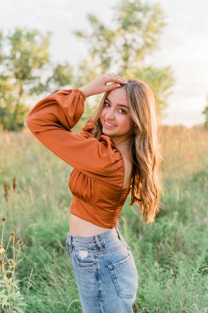 A person in a brown top and jeans poses playfully in a grassy meadow at sunset.