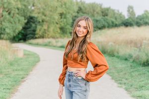 A person in an orange sweater and jeans poses outdoors in a natural setting during autumn.