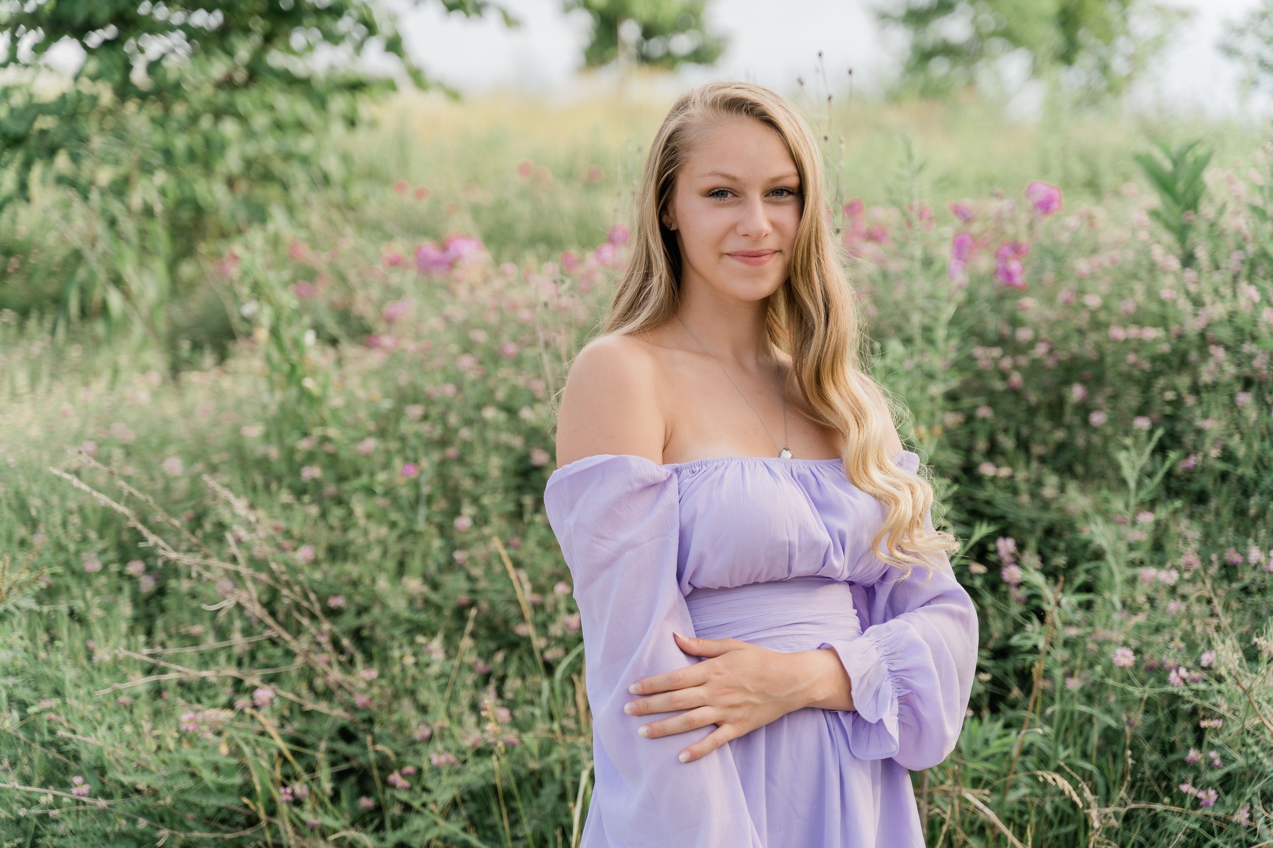A woman in a lavender dress poses gracefully in a garden setting with blooming flowers and greenery.
