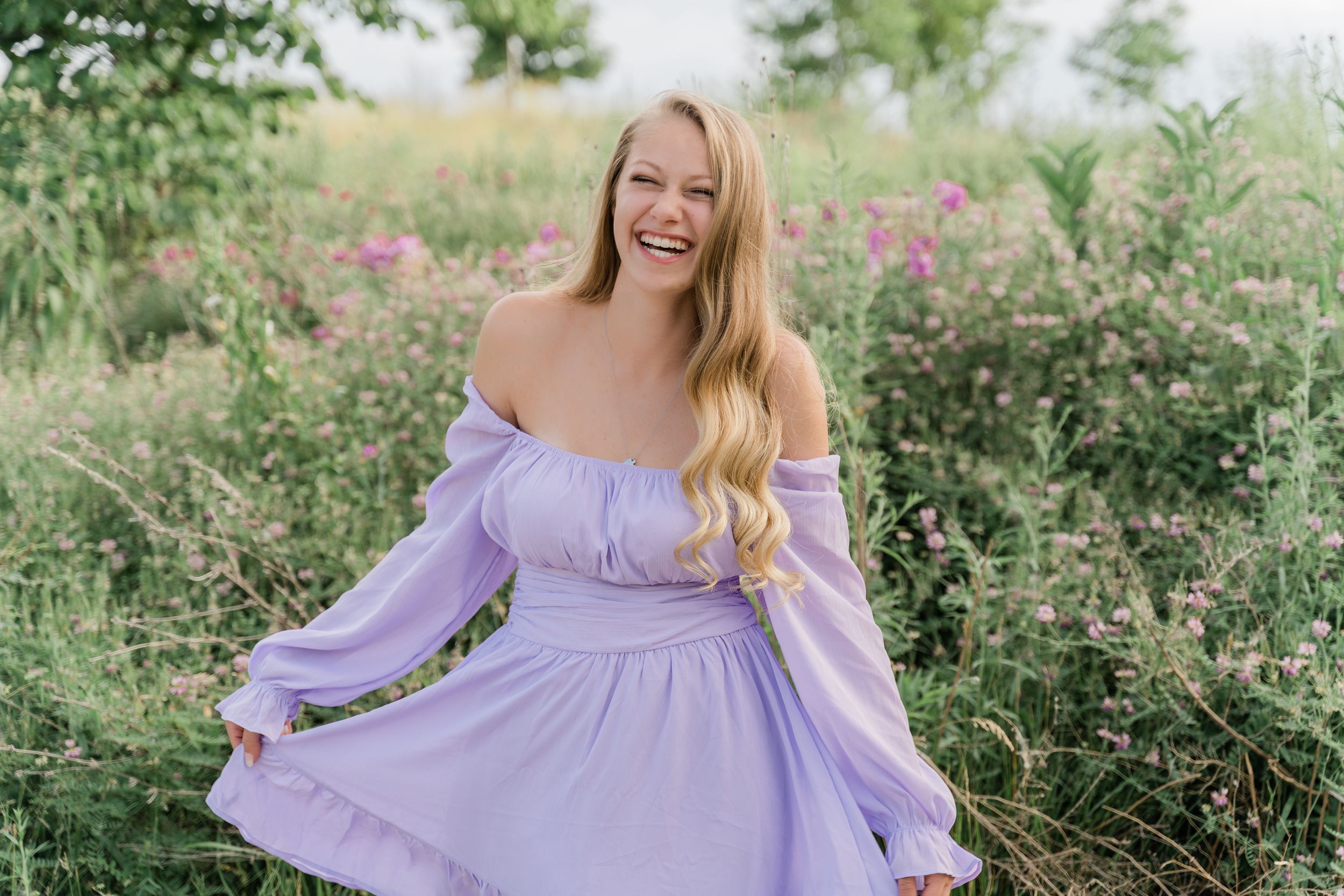 A person in a flowing lavender dress twirls gracefully in a garden filled with pink wildflowers and green foliage.