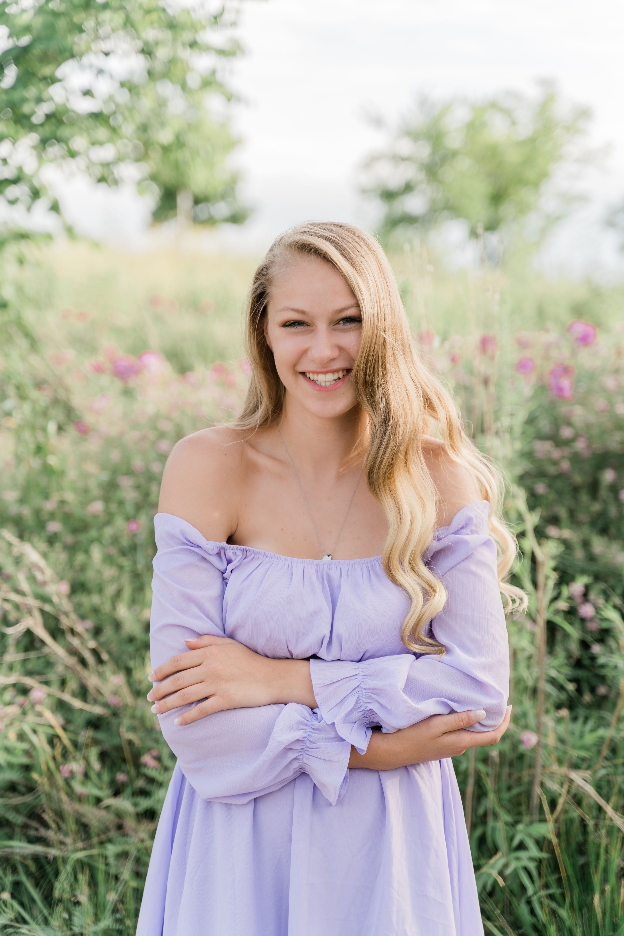 A person in a light purple off-shoulder dress poses outdoors against a backdrop of flowering bushes and greenery.