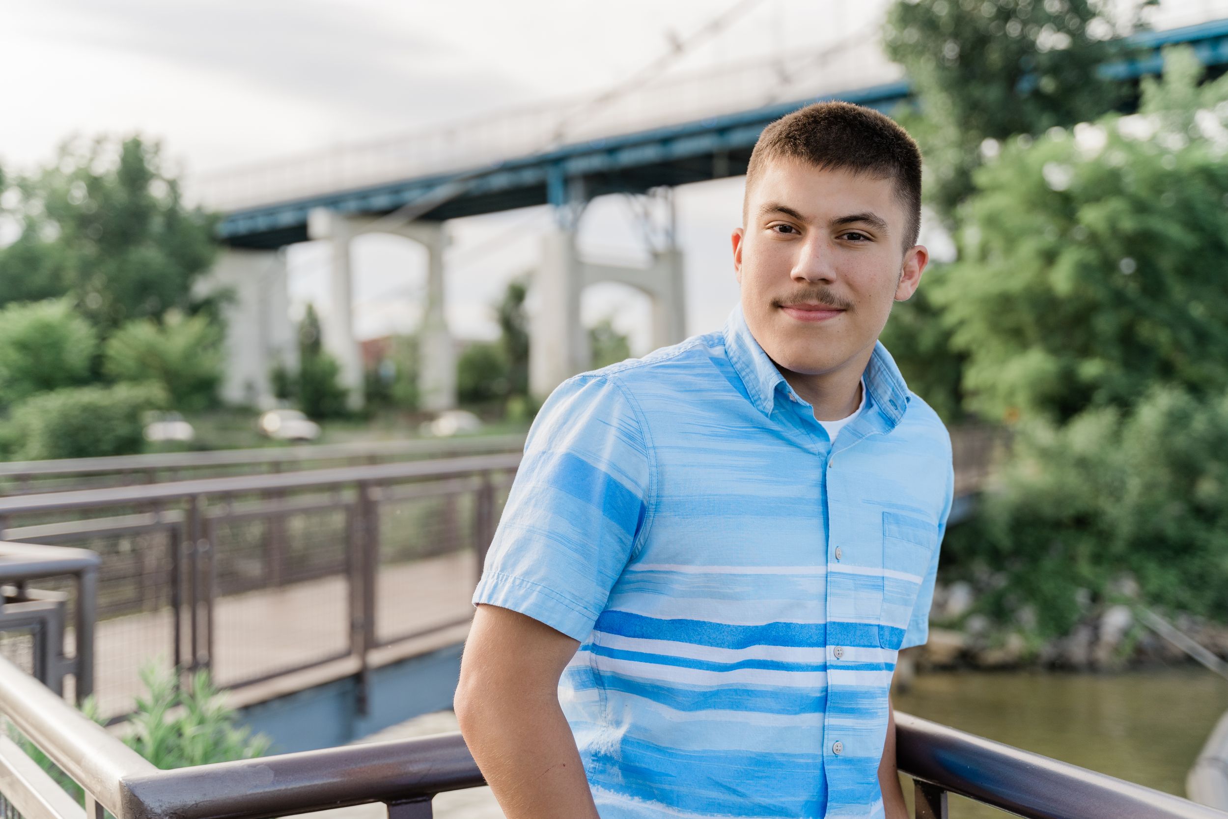 Person in blue and white striped polo shirt stands by metal railing with bridge and river scenery behind.
