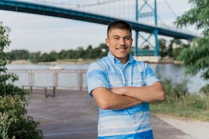 A person in a blue plaid shirt stands smiling with arms crossed near a waterfront bridge.