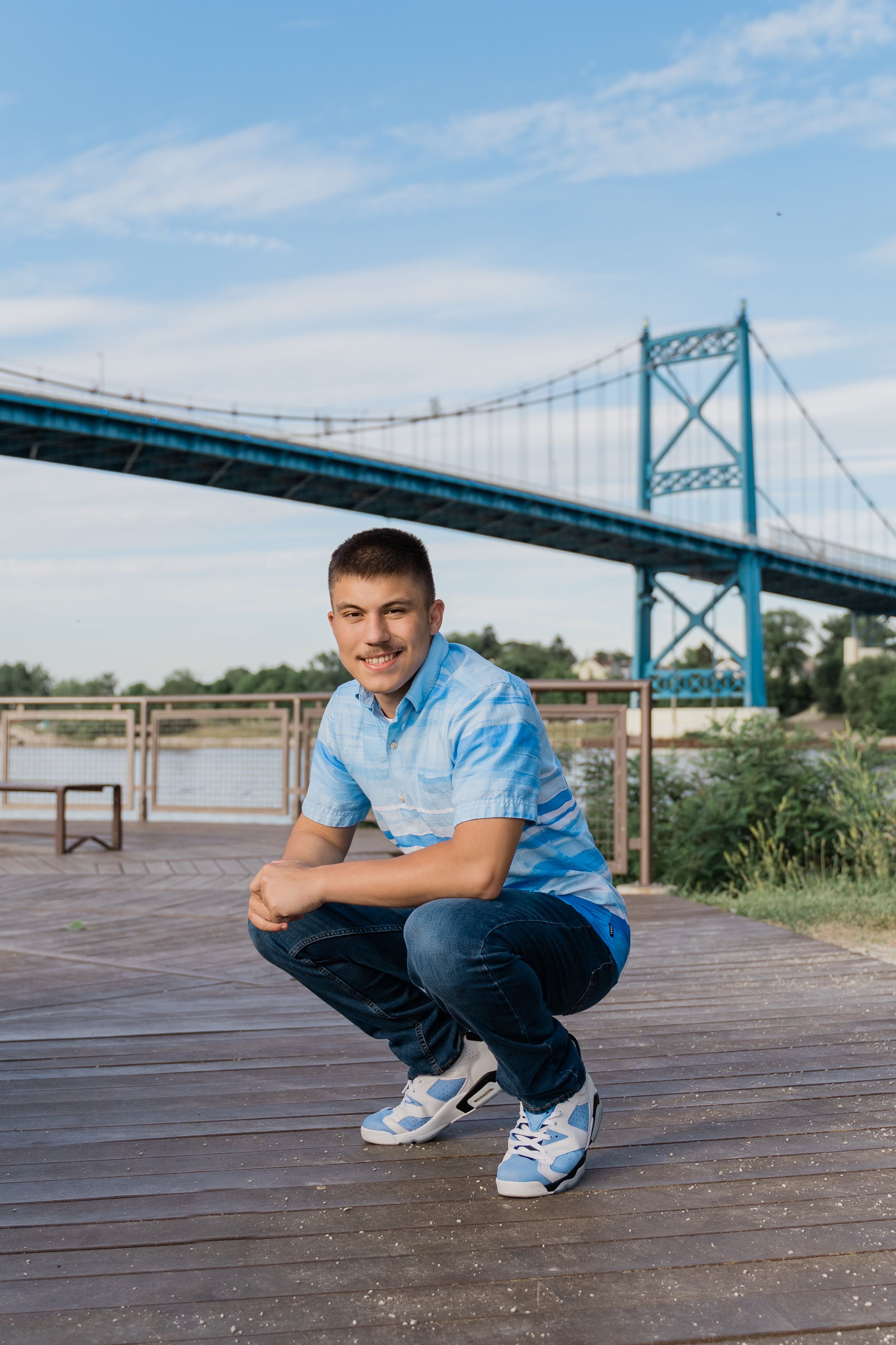 Person in light blue shirt and jeans crouches on wooden boardwalk with Ambassador Bridge visible in background.