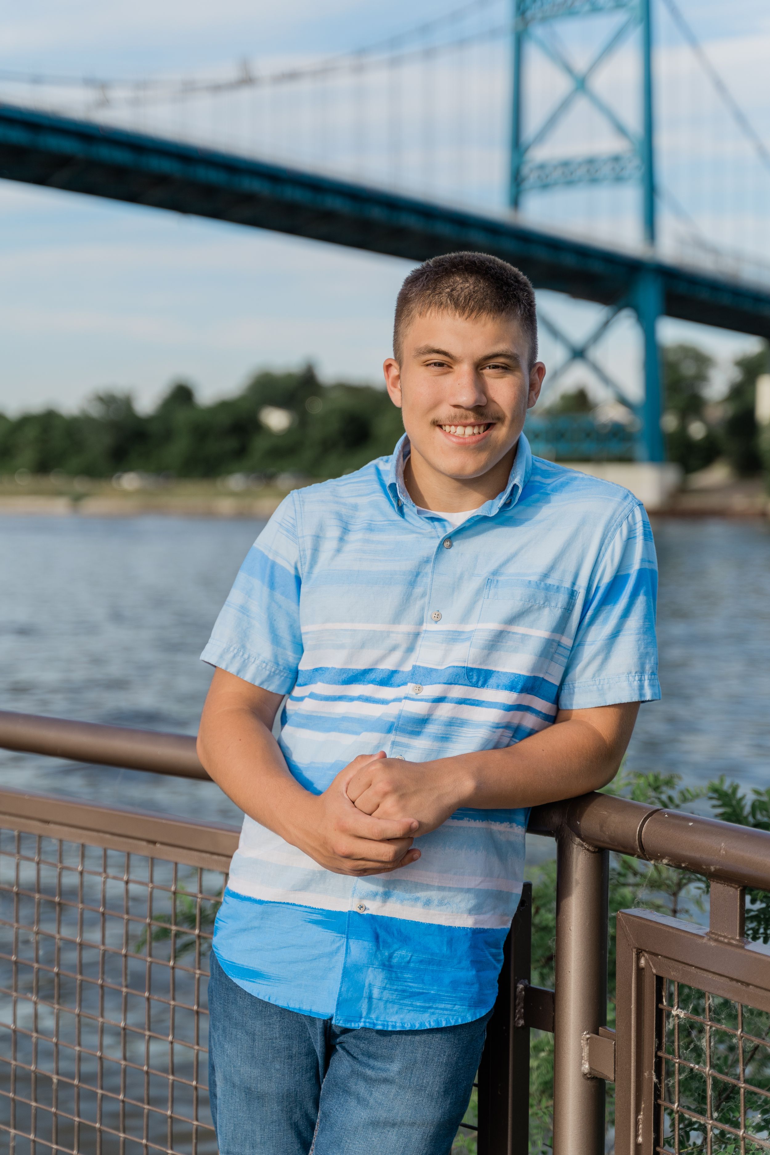 Person in light blue striped shirt poses near waterfront with large bridge structure visible in background.