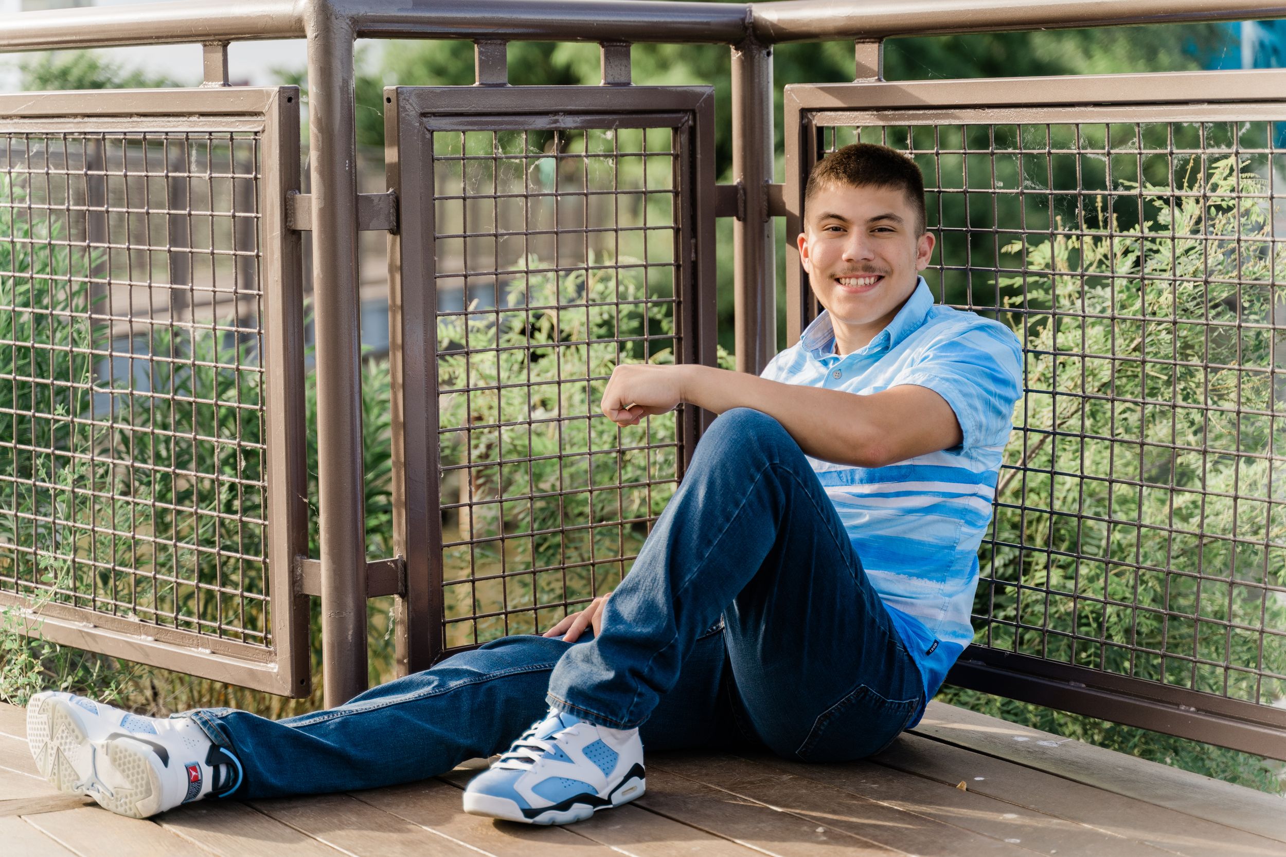 Someone in a light blue polo shirt and jeans sits casually on a wooden deck with wire mesh fencing in the background.
