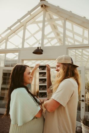 Couple smiles at each other while holding ultrasound photos in front of glass greenhouse at golden hour.