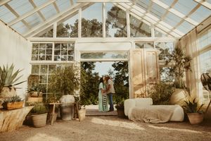 Expecting couple embraces in a bright, plant-filled greenhouse with large glass windows and cozy decor.