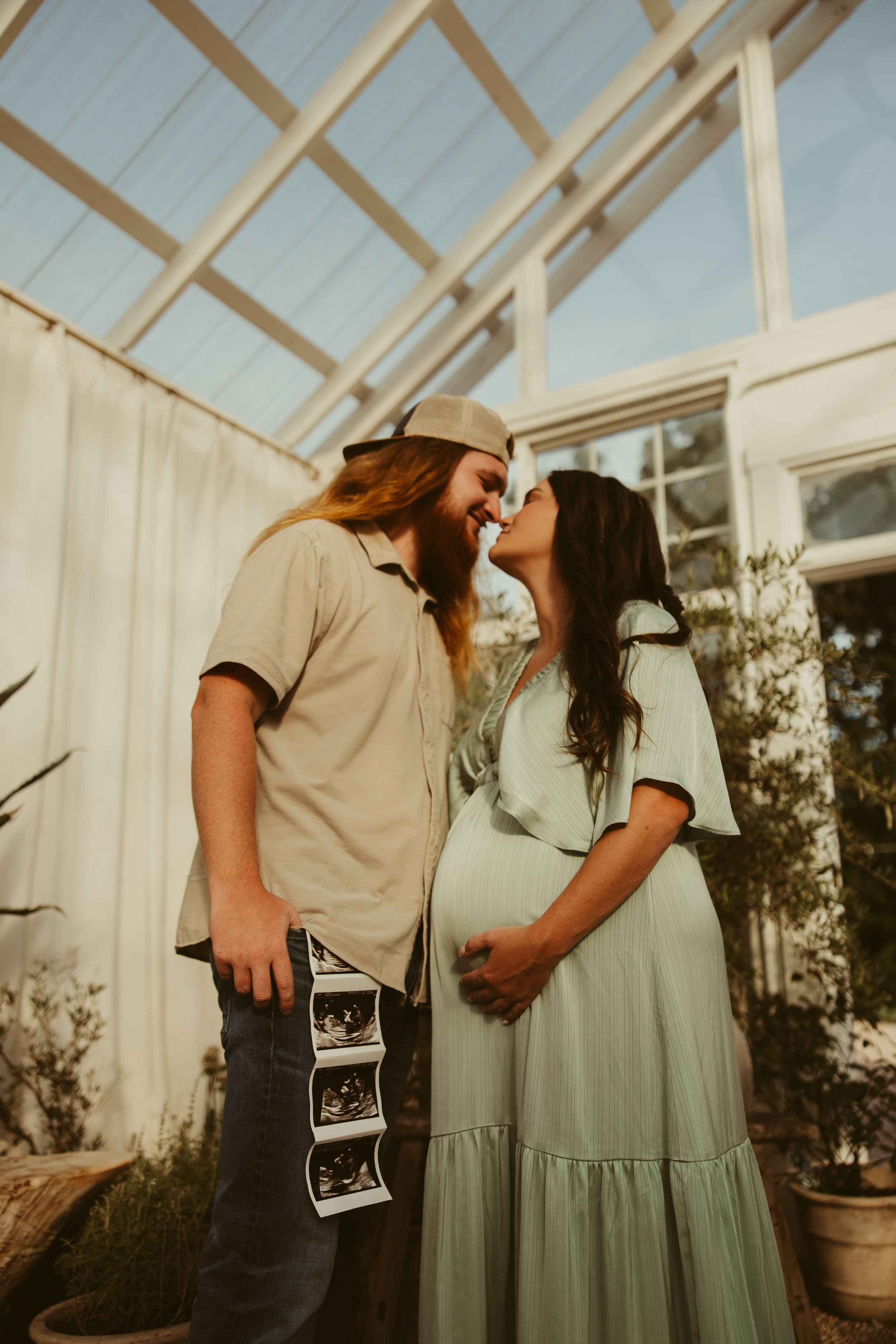 Smiling couple touches foreheads inside a greenhouse, man holding ultrasound strip beside pregnant partner.