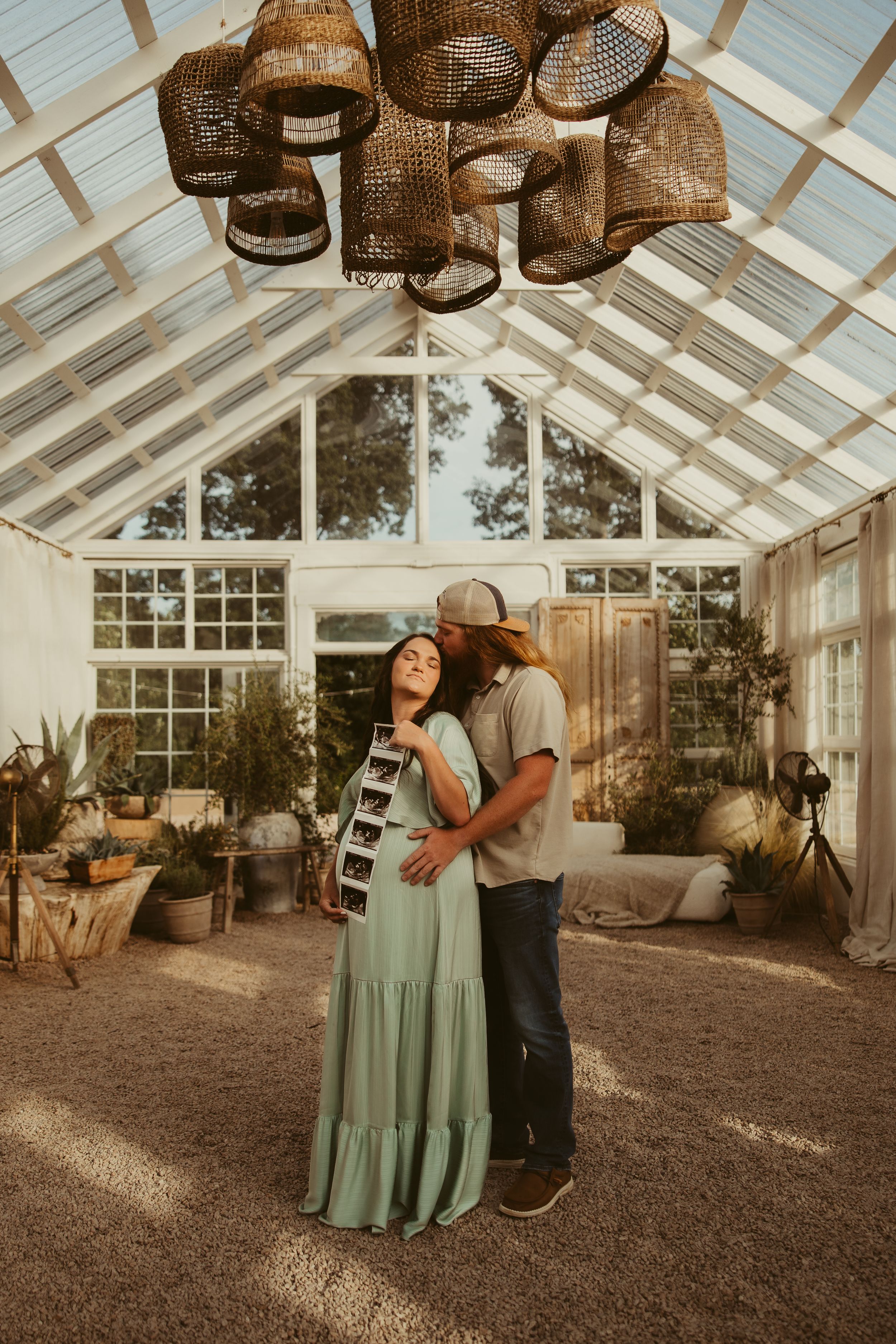 A couple shares a tender moment in a sunlit greenhouse, holding ultrasound photos beneath woven pendant lights at Fleurish Greenhouse.