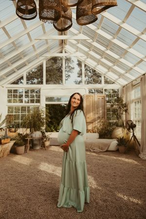 A smiling pregnant woman in a mint green maxi dress stands in a bright, plant-filled glass greenhouse in Taylorsville, NC.