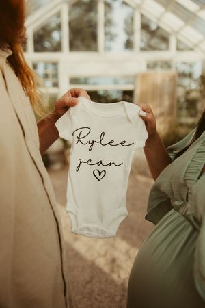 Two women hold a white baby onesie reading Rylee Jean with a heart in a greenhouse setting.