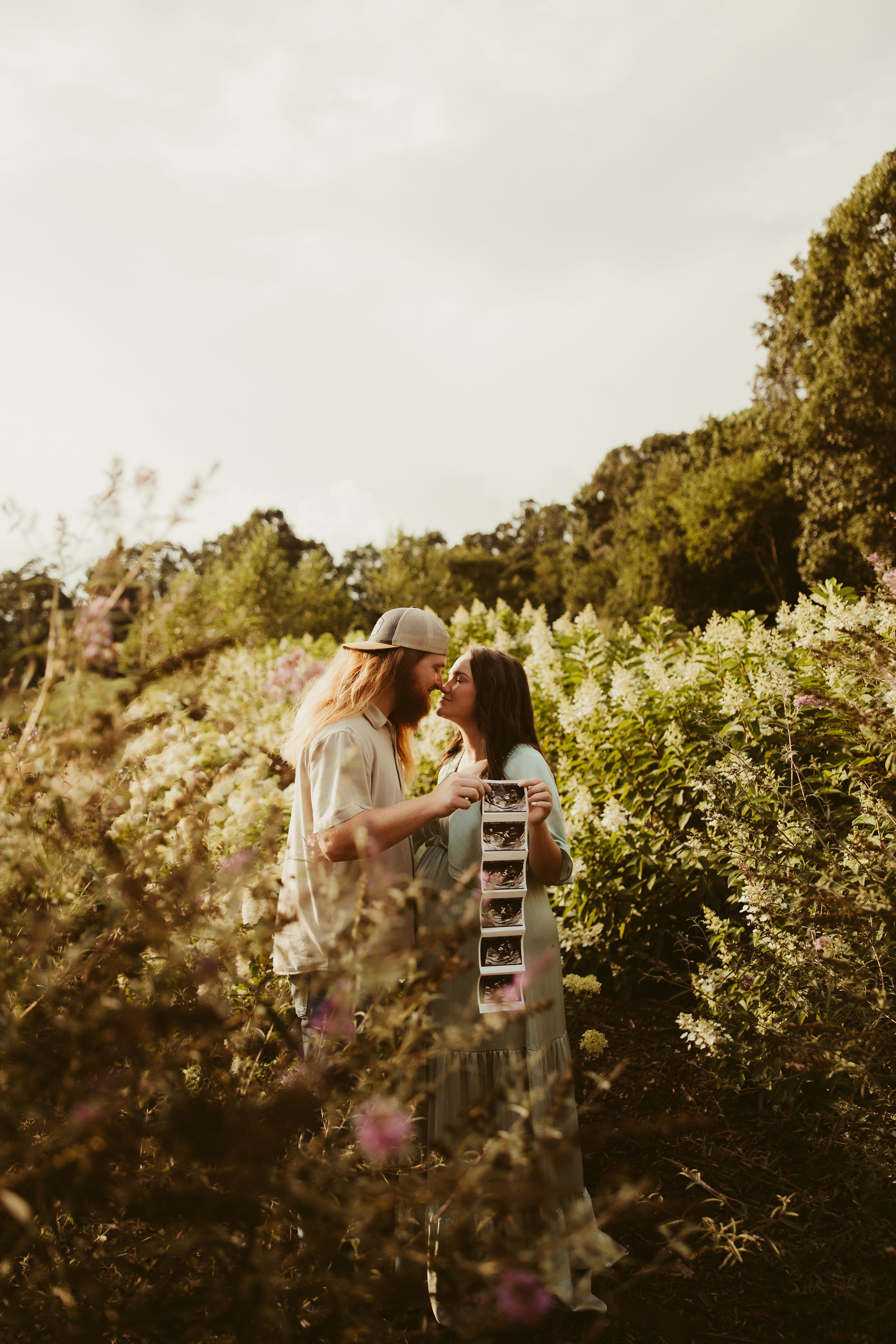 A couple shares a romantic kiss in a lush garden while holding ultrasound photos on a cloudy day.
