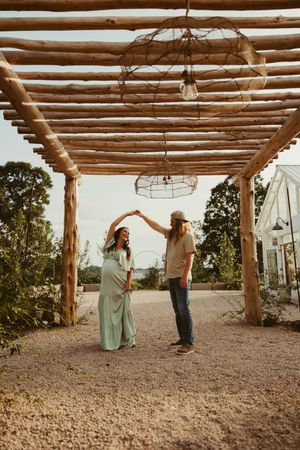 Expecting couple dances joyfully under a rustic wooden pergola with hanging lights outdoors.