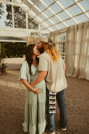 Couple shares a tender kiss inside a sunlit greenhouse, man holding ultrasound photos behind his back.
