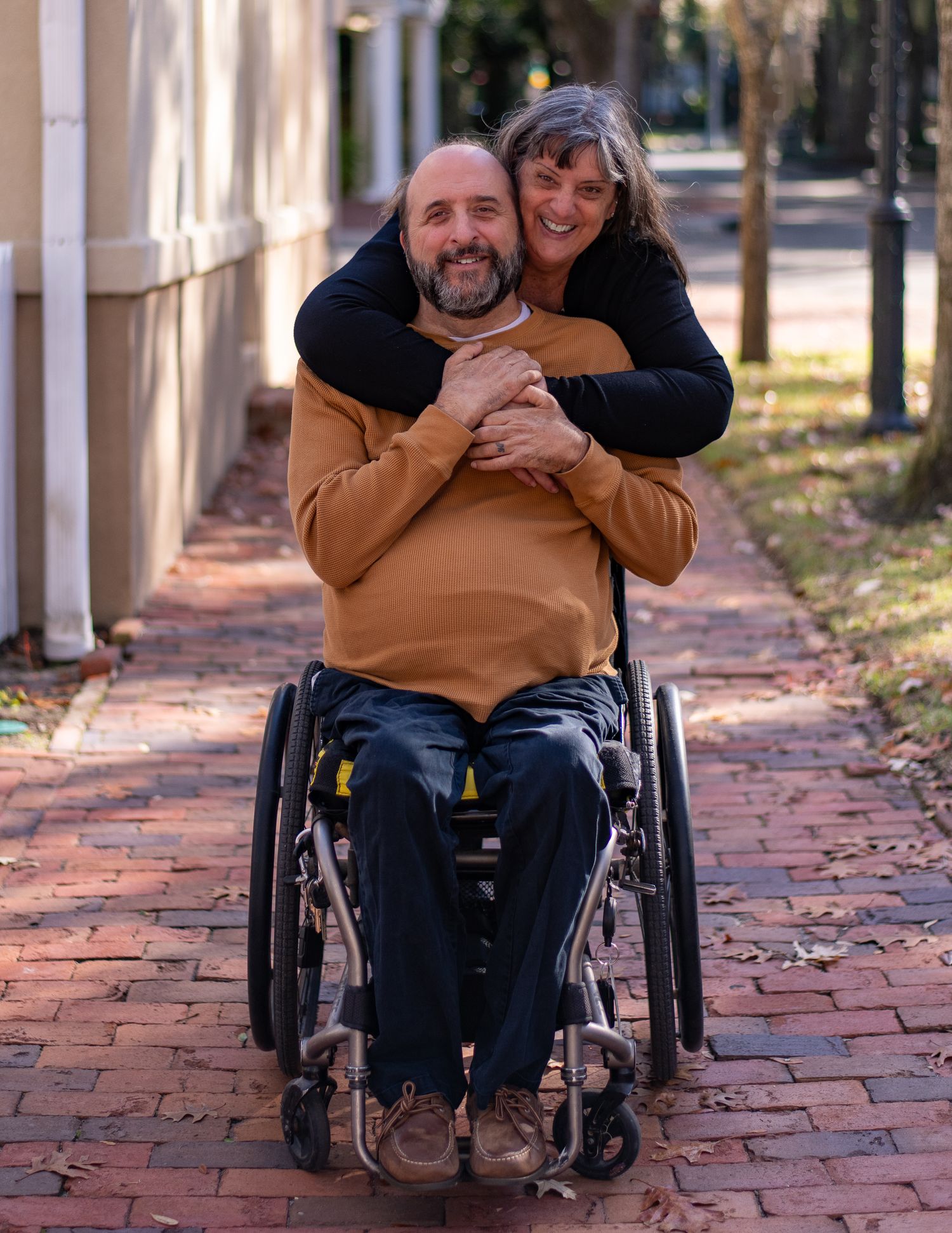 A couple shares a joyful moment on a brick walkway lined with columns, with one person seated in a wheelchair.