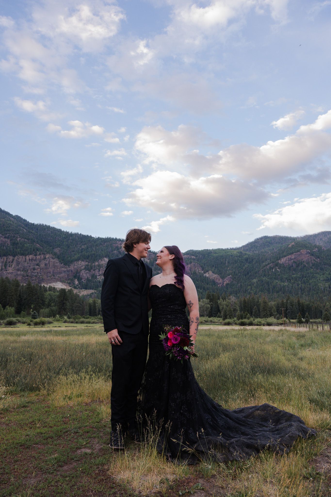 Couple in matching black wedding attire standing in mountain meadow under blue sky.