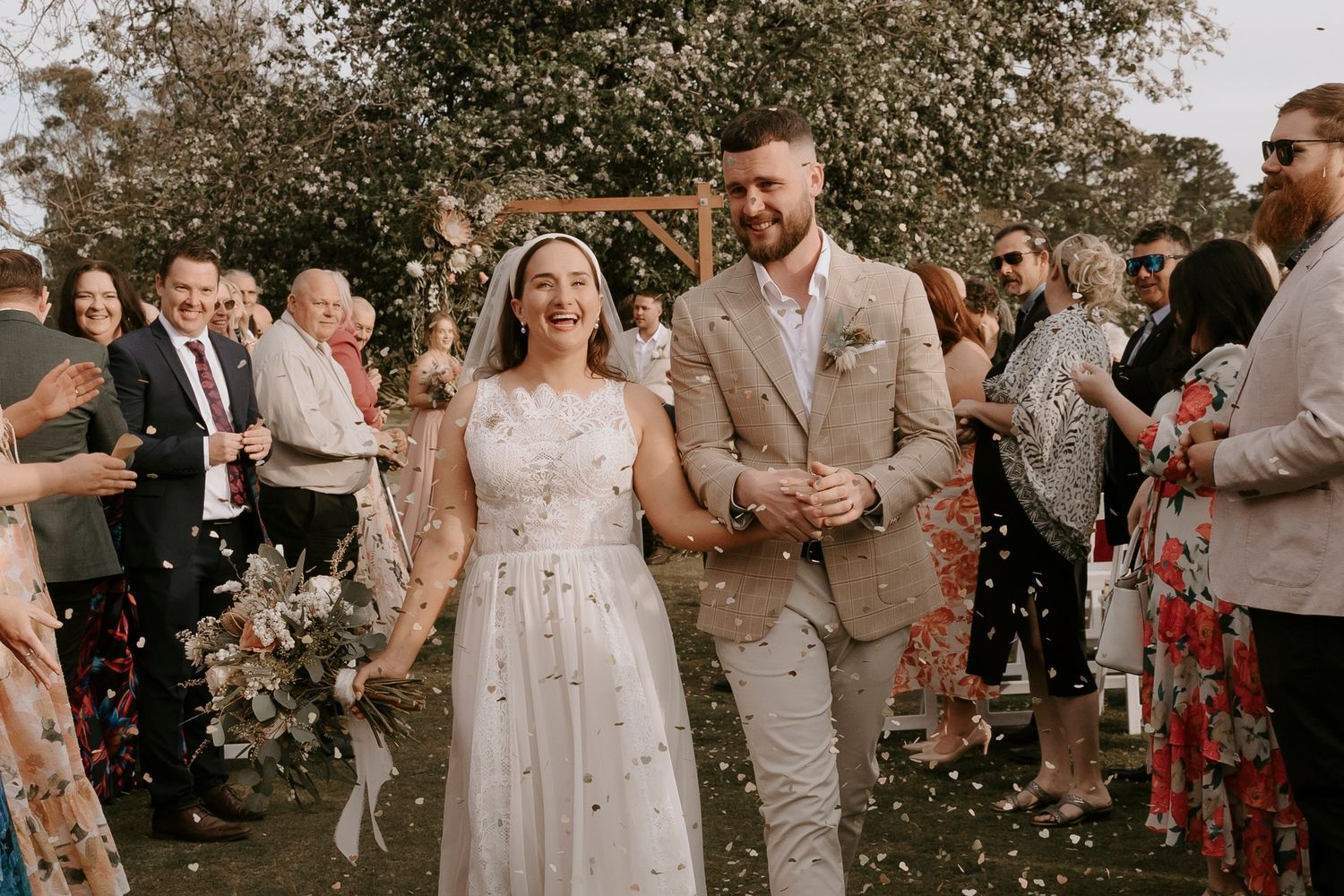 Wedding guests gather outdoors for a celebration with soft natural lighting and floral decorations in the background.