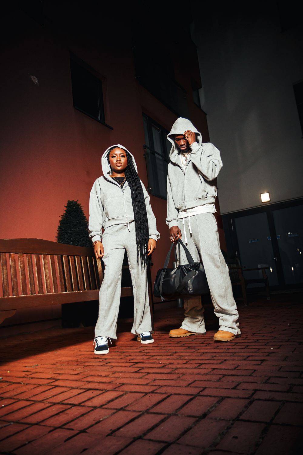 Two people in matching light gray sweatsuit outfits pose on brick pavement at night under dramatic lighting.