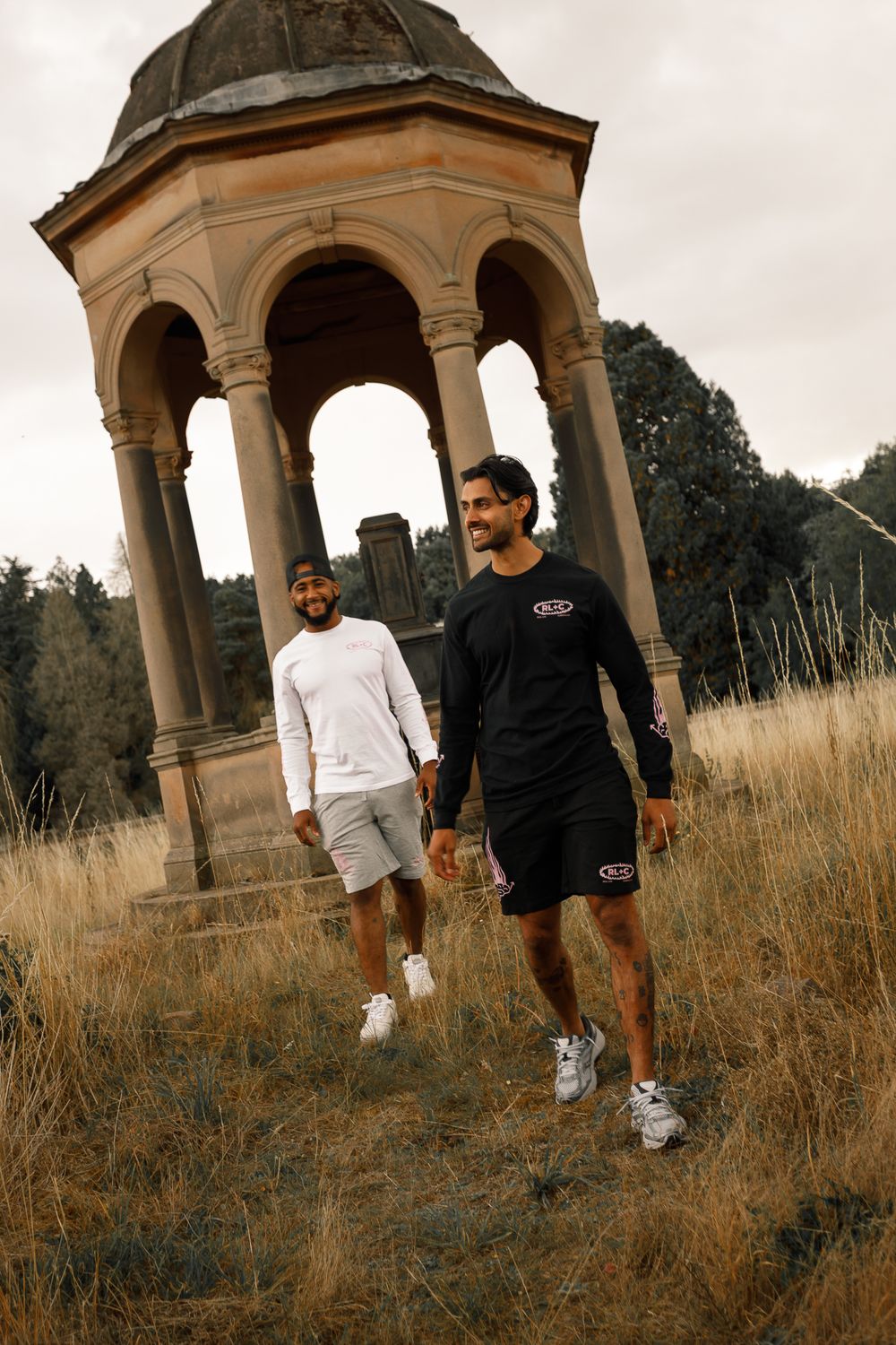 Two people walking near a stone gazebo structure with arches in a field of tall grass on an overcast day.