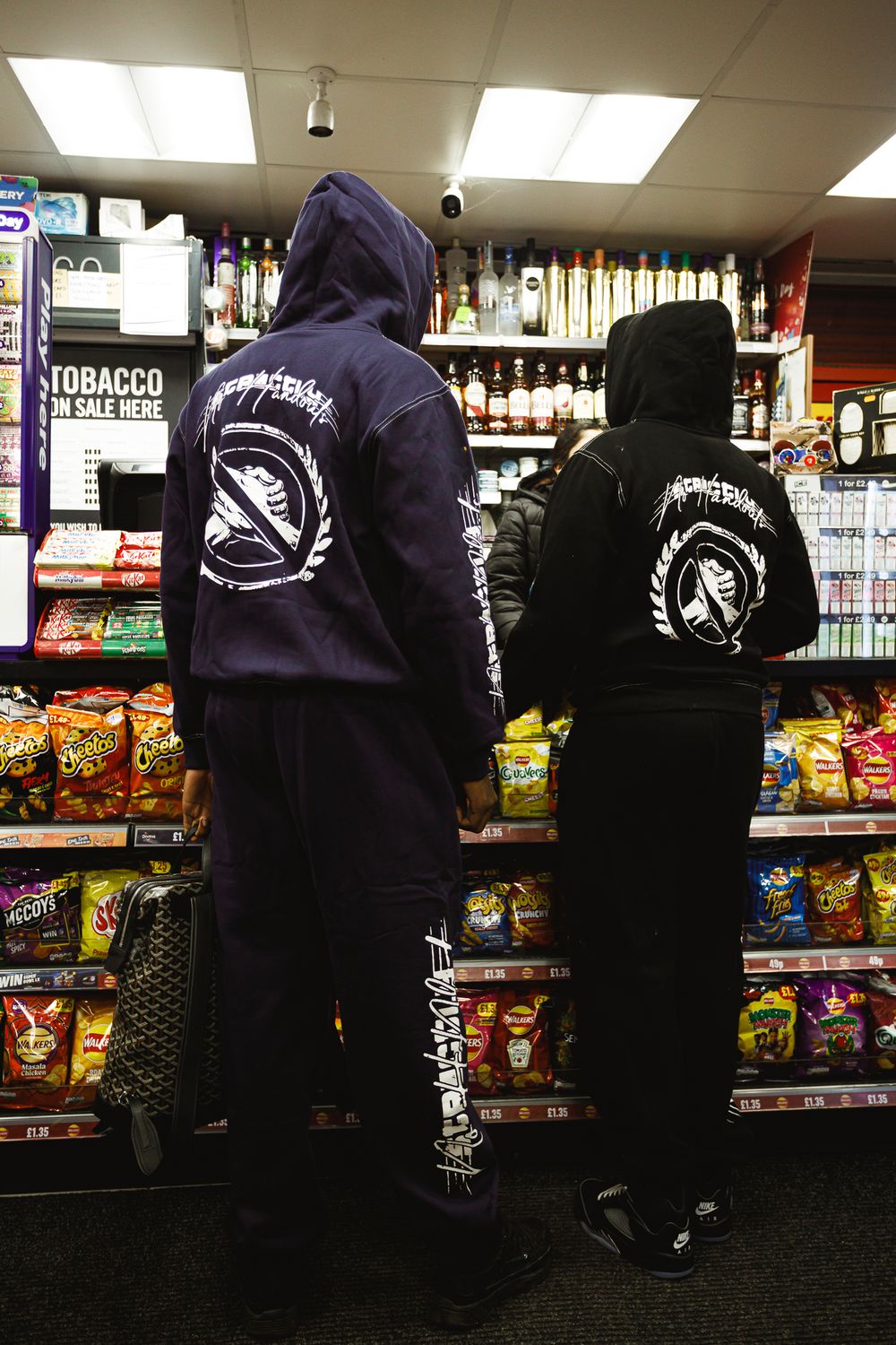 Two people wearing black and navy hooded sweatshirts with matching designs stand in front of a convenience store snack aisle.