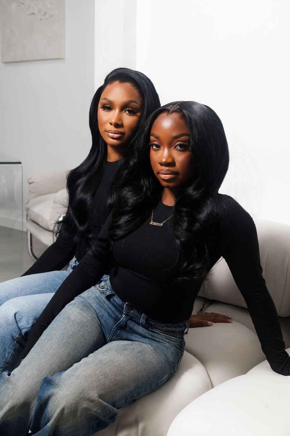 Two women in matching black sweaters and blue jeans pose together on a white couch in a minimalist setting.
