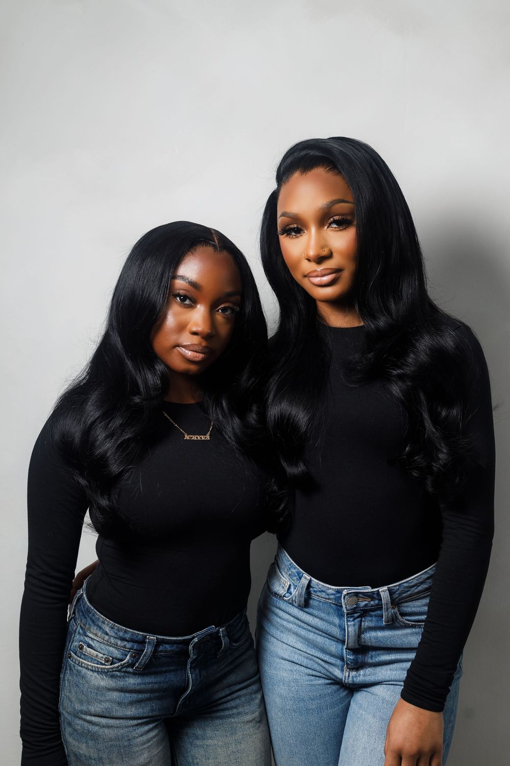 Two women wearing black long sleeve tops and blue jeans pose together against a light gray background.