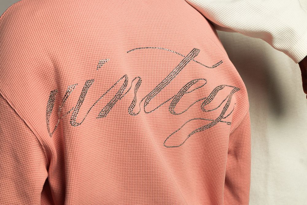 Close-up detail of a peach-colored sweatshirt with 'vinteg' script lettering embroidered on it.