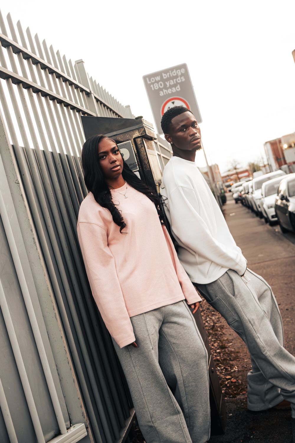 Two people pose casually against a metal fence wearing coordinated casual streetwear outfits.