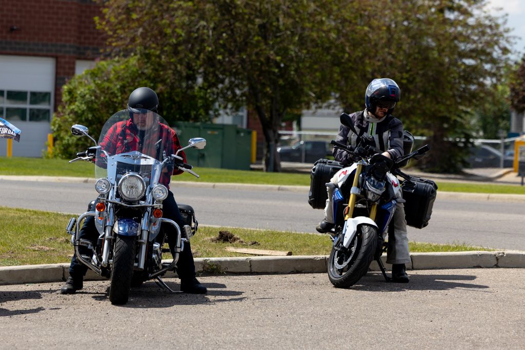 A line of parked motorcycles on a street with brick buildings and trees in the background.
