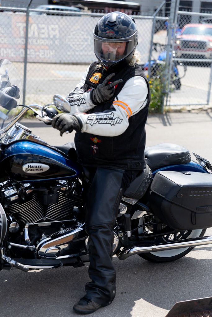 A motorcycle rider in black gear sits on a parked blue and black cruiser bike in a parking lot.