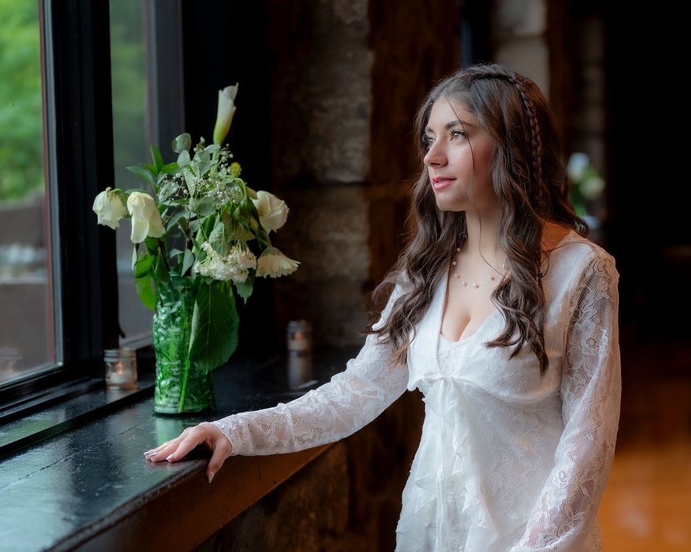 Woman in white lace dress stands by window with green vase of white roses in romantic indoor setting.