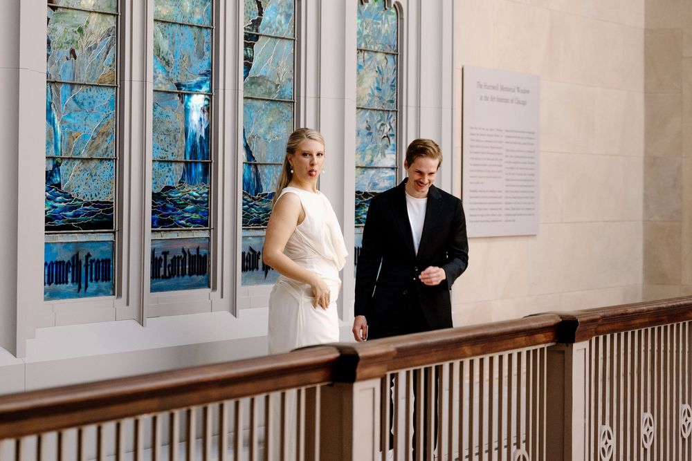 A couple in formal attire stands together in an elegant hallway with stained glass windows.