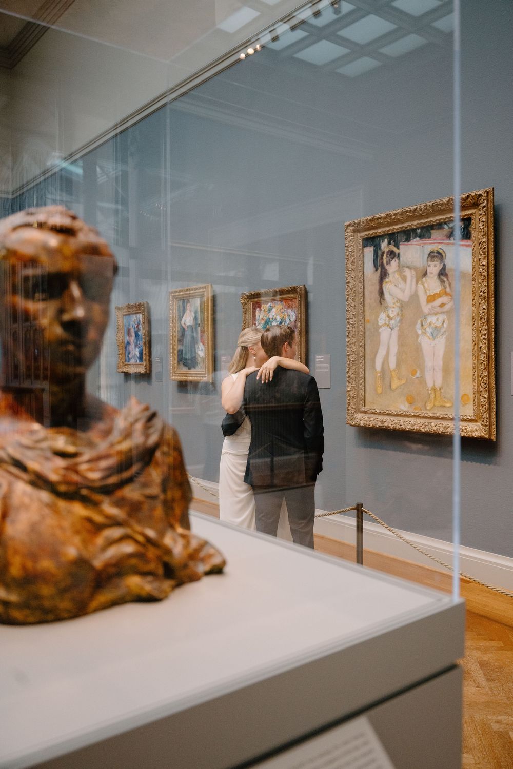Art gallery interior showing framed paintings mounted on gray walls with display pedestals in foreground.